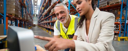 With a background of a large warehouse, one person in business attire points to a computer screen as another person in a hi-vis vest watches.